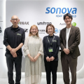 ソノヴァ・ジャパン株式会社のオフィスにて、「Sonova HEAR THE WORLD」のロゴや傘下ブランド(unitron、Sennheiser、Audiofona等)のロゴが並ぶ白い壁の前に、ソノヴァ・ジャパンとオースタンスの担当者4名が横一列に並んで笑顔で写っている。左端と中央右の2名はソノヴァのロゴ入りポロシャツを着用。壁面左側にはヘッドフォンのディスプレイが見える。