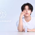 「hada to kokoro(肌と心)」のブランドアンバサダーであるJO1・河野純喜氏を起用したブランドビジュアル。淡いブルーグラデーションの背景の前で、白いTシャツを着た河野純喜氏がテーブルに腕をつき、ブランドのオールインワンクリーム化粧水を手前に置いた状態でカメラを真っ直ぐ見つめている。左側にはブランドロゴマークと「hada to kokoro°」のロゴテキストが配置されている。