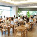 「Oisix Retail ADs」の社食・学食リアルプロモーションメディアのイメージ写真。木製のテーブルと椅子が並ぶ明るく開放的な職域食堂で、ビジネスカジュアルの男女のグループが食事をしながら談笑している様子。大きな窓から緑の木々が見え、観葉植物も配置された清潔感のある空間で、日常的に多くの人が利用する社食の雰囲気を表現している。