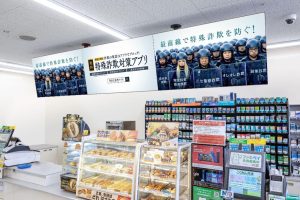 ファミマTVの店内サイネージで放映される特殊詐欺対策アプリの啓発広告。コンビニのレジ上部に設置された横長ディスプレイに「最前線で特殊詐欺を防ぐ！」の広告が表示されている
