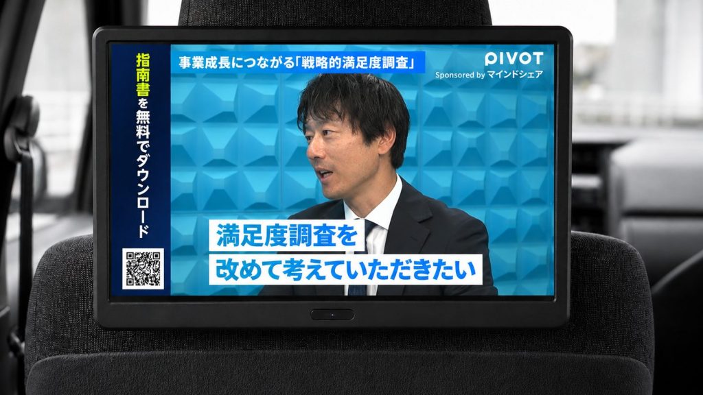 タクシー車内モニターに映るPIVOT番組で、マインドシェアの出演者が「満足度調査を改めて考えていただきたい」と語る場面。左側に指南書無料ダウンロードのQRコードを表示