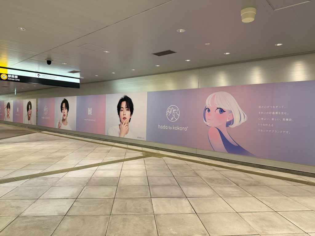 渋谷駅東口地下広場B1F 東棟連絡通路に掲出された「hada to kokoro(肌と心)」の巨大壁面広告の全景。ピンクから薄紫のグラデーション背景に、ブランドアンバサダーの河野純喜氏(JO1)のポートレートが複数並んで展開されており、右端にはブランドロゴとイラストキャラクター、「肌と心がつながって、きれいの好循環を生む。心地よい、楽しい、高機能、3つを叶えるスキンケアブランドです。」というブランドコピーが記載されている。銀座線の案内サインが天井に見える。